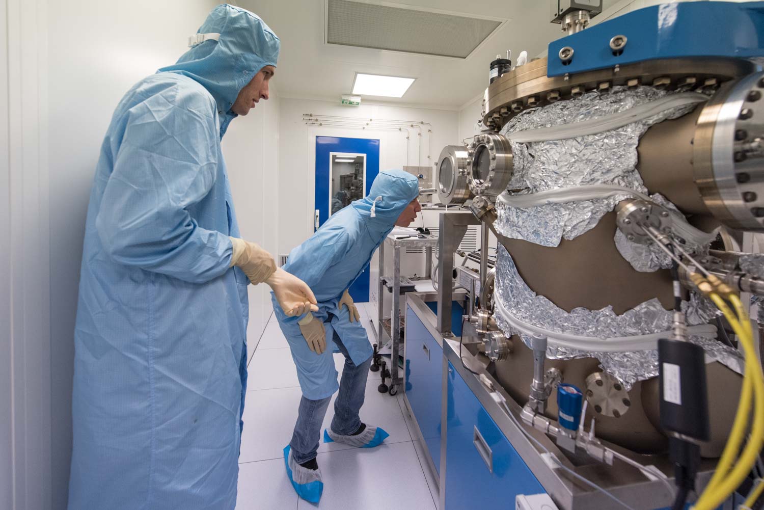 ENS, Département de physique, LPA. Laboratoire Pierre Aigrin . recherche sur les circuits quantiques hybrides. Takis Kontos et un de ses doctorants Tino Cubaynes dans la salle Blanche du département de physique observent à travers les hublots de cette chambre à ultra-haut vide un échantillon de circuit électronique sur lequel sont en train d'être déposés de fines couches de métaux rares. Ces circuits, sorte de "boites à particules" sont utlisées pour piéger les éléctrons ou les photons dont les propriétés quantiques sont étudiées par les chercheurs du laboratoire. La pureté des échantillons est une condition indispensable au fonctionnement des expériences à cette échelle infra nanométrique. C'est pourquoi ils sont concus en salle blanche sous ultra vide.
